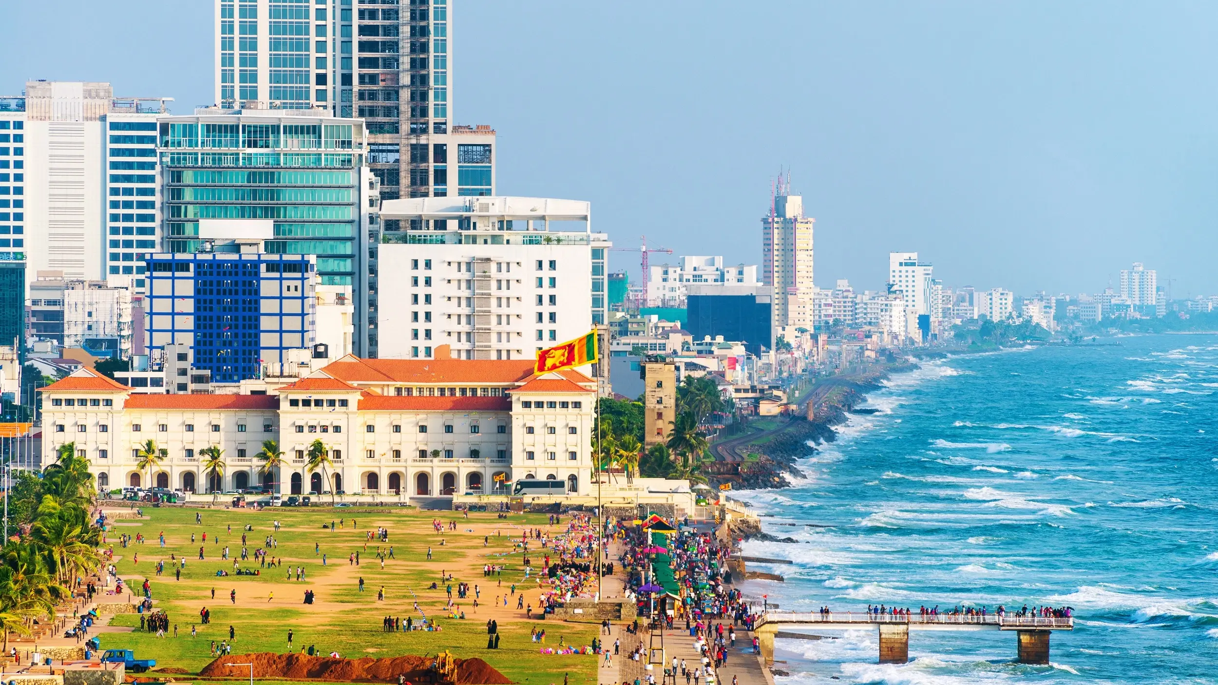 People gathered at a park and along the building-lined shoreline with a jetty and waves coming into shore, at Galle Face Green, Colombo. Image credit: stock.adobe.com