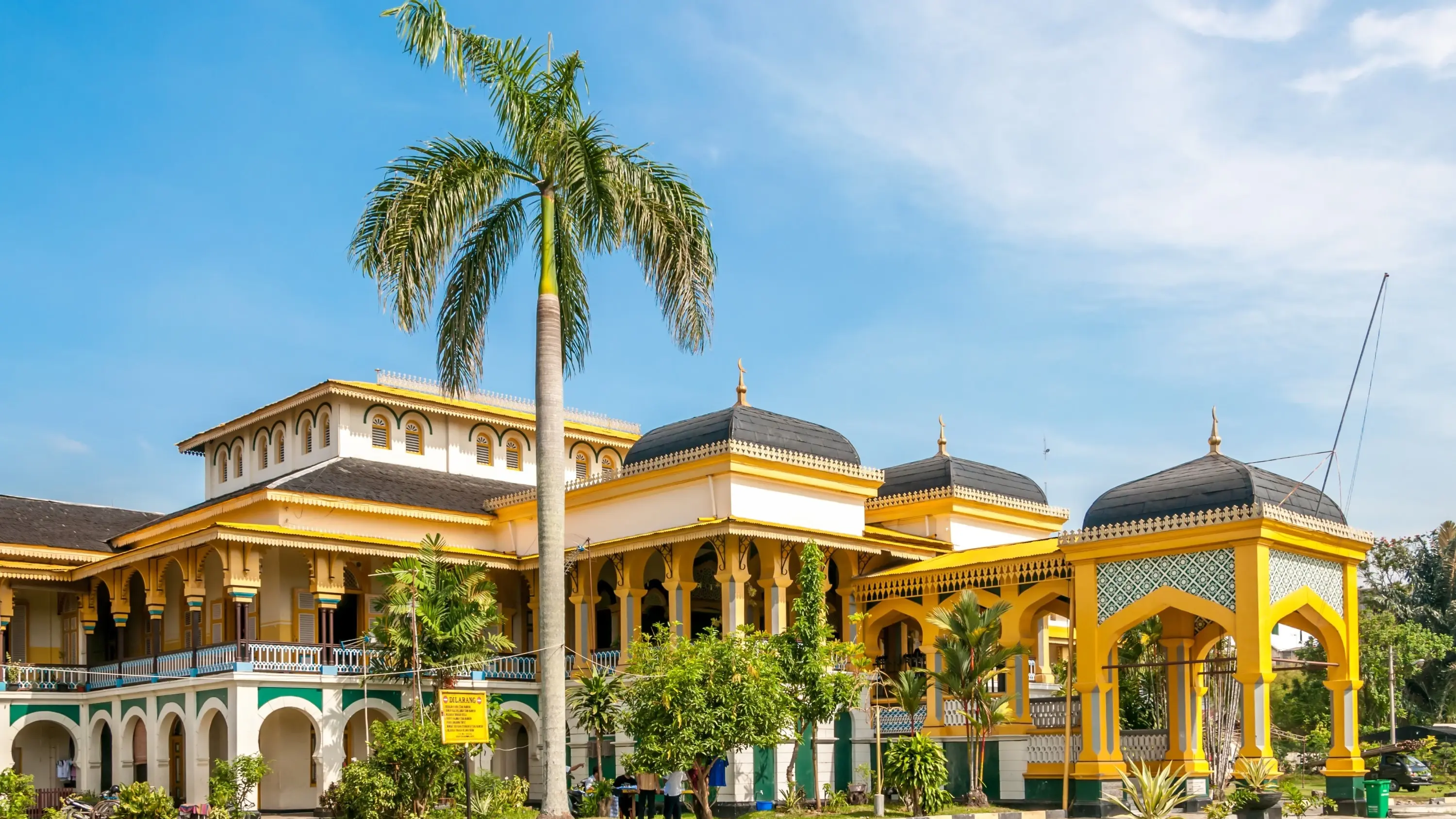 The ornate, bright-yellow Maimun Palace with palm trees out front, Medan, Indonesia. Image credit: stock.adobe.com