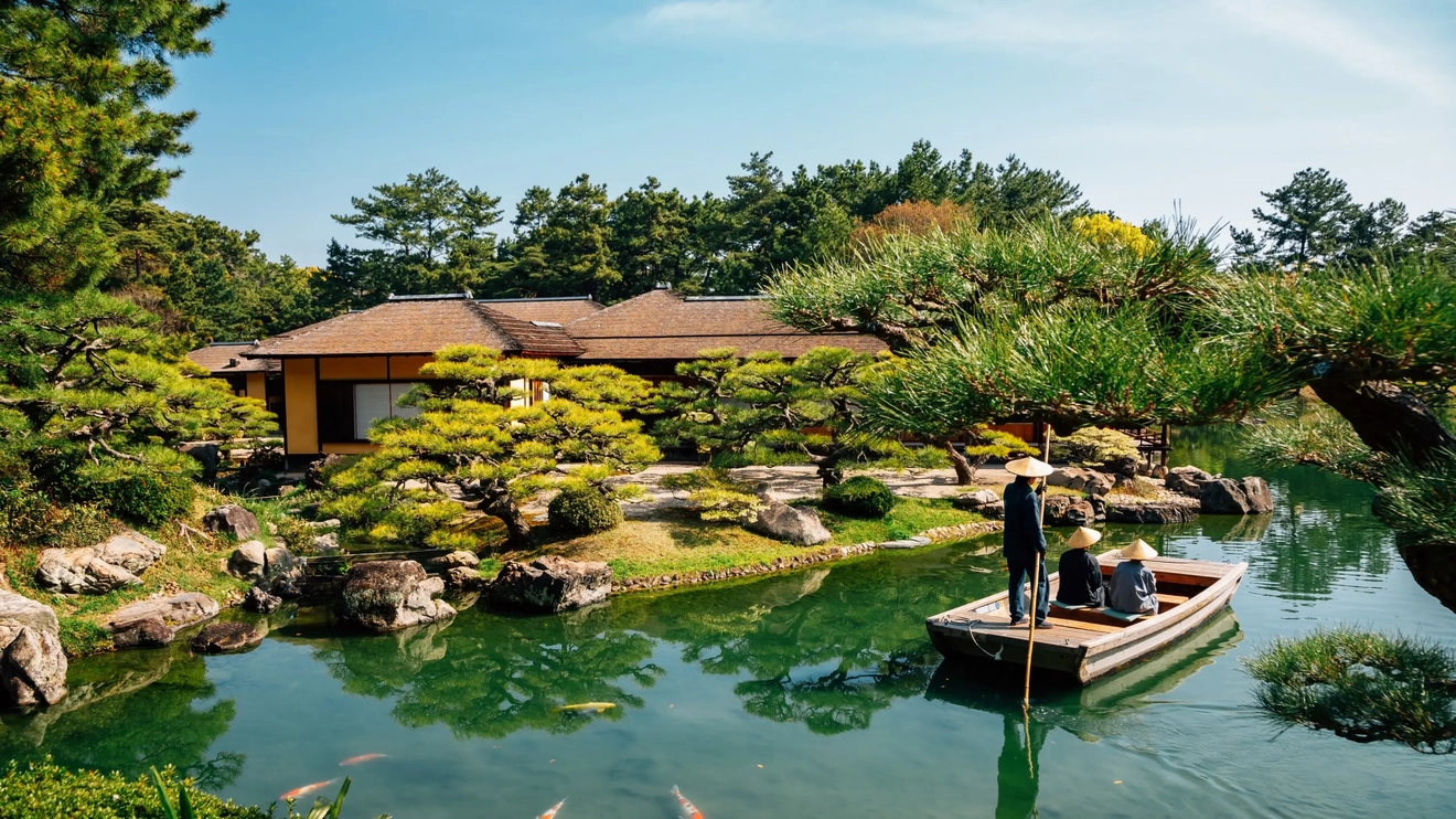 Three people on a wooden boat in a stream with carp in the foreground and a traditional building and trees in the background, at Ritsurin Park, Takamatsu, Japan. Image credit: stock.adobe.com
