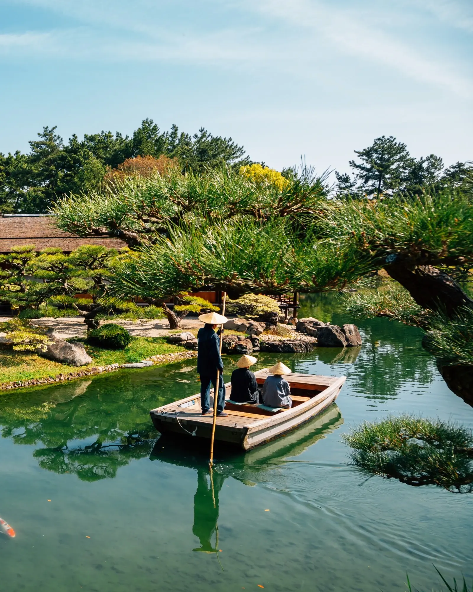 Three people on a wooden boat in a stream with carp in the foreground and a traditional building and trees in the background, at Ritsurin Park, Takamatsu, Japan. Image credit: stock.adobe.com