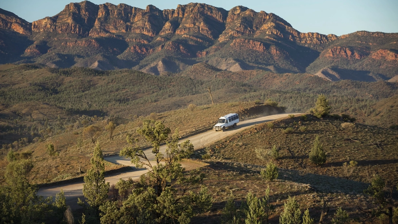SUV driving down outback road, Flinders Ranges in background. Learn about the ancient Aboriginal history of this area on a Gorgeous Gorges tour. Image credit: Tourism Australia.
