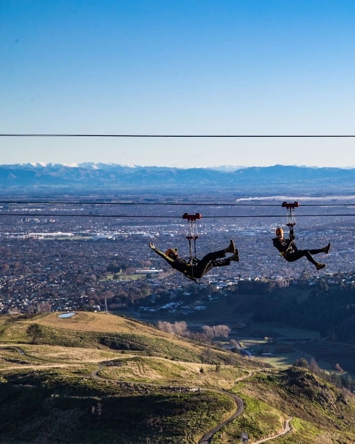 Two people ziplining at Christchurch Adventure Park with Christchurch and mountains in background, Christchurch, New Zealand. Image credit: ChristchurchNZ