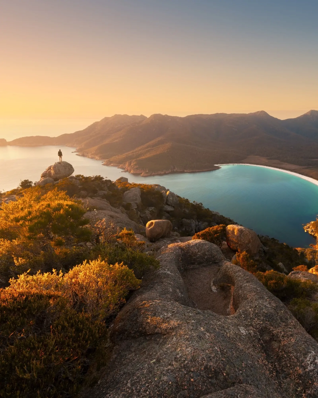 Person standing on boulder overlooking Wineglass Bay in Freycinet National Park at sunset. Image credit: Matthew Donovan/Tourism Tasmania