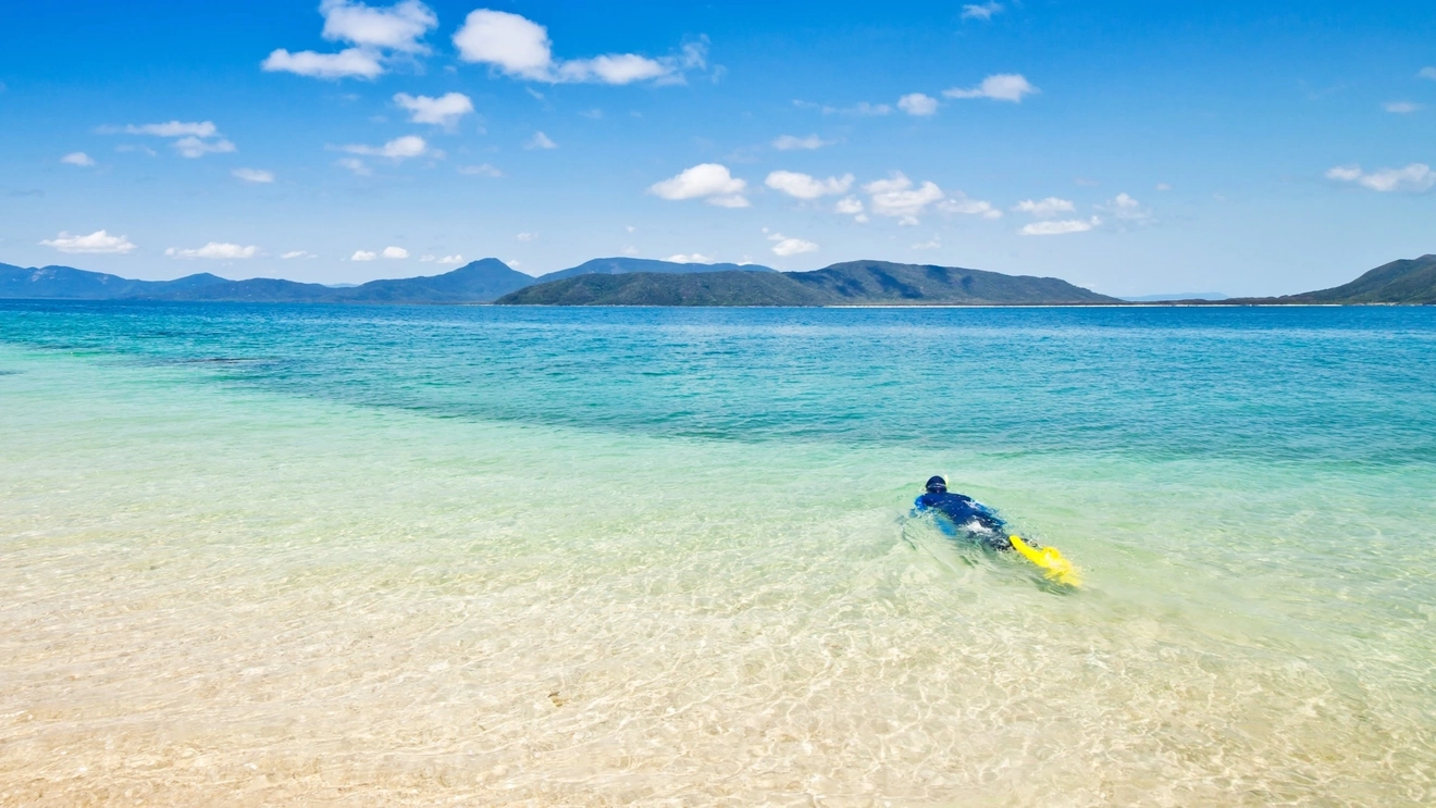 Person snorkelling in clear, turquoise water off Nudey Beach, Fitzroy Island, with islands in the background.
