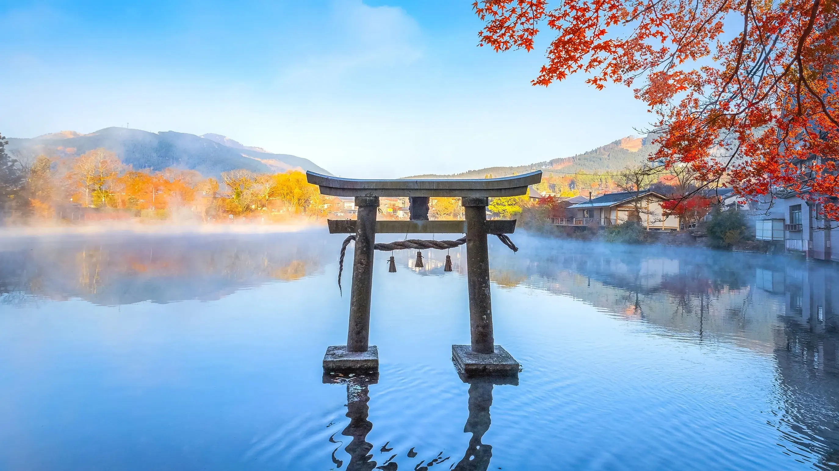 Torii temple gate in the reflective Lake Kinrinko in Yufuin, Oita with a blue sky and trees with autumn leaves. Image credit: stock.adobe.com