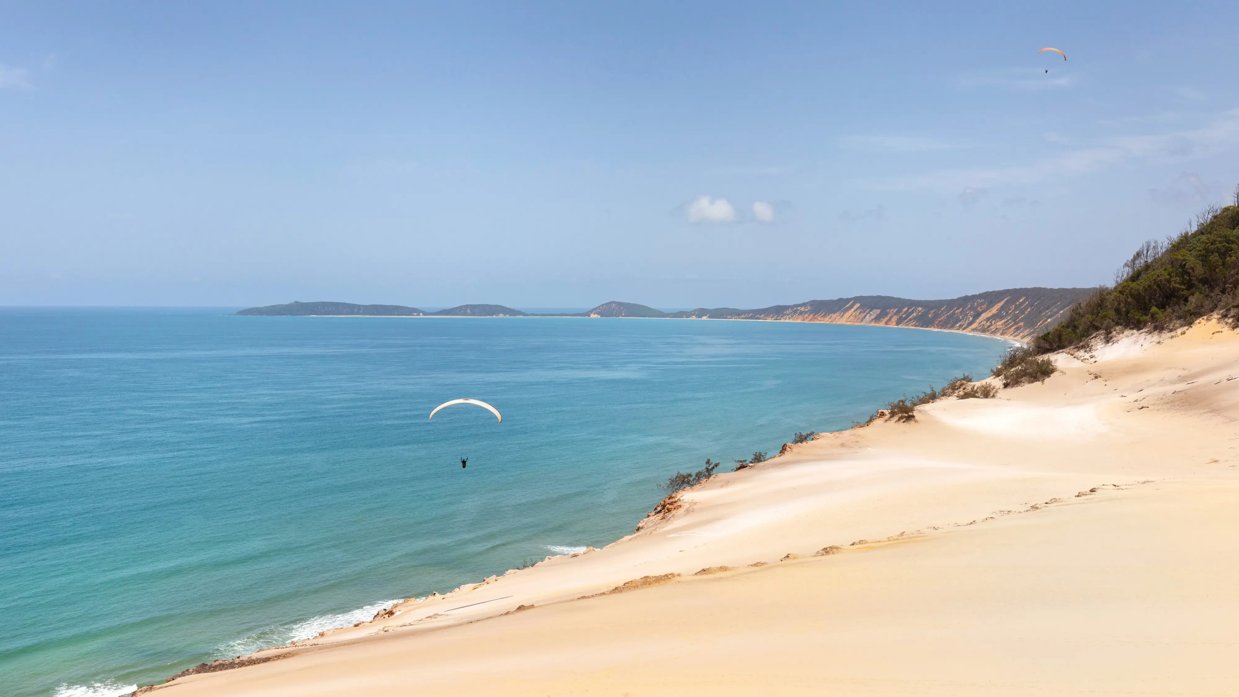 Aerial shot of Rainbow Beach, with bright blue water, gold sand, pink sand and a lone paraglider. Image credit: stock.adobe.com