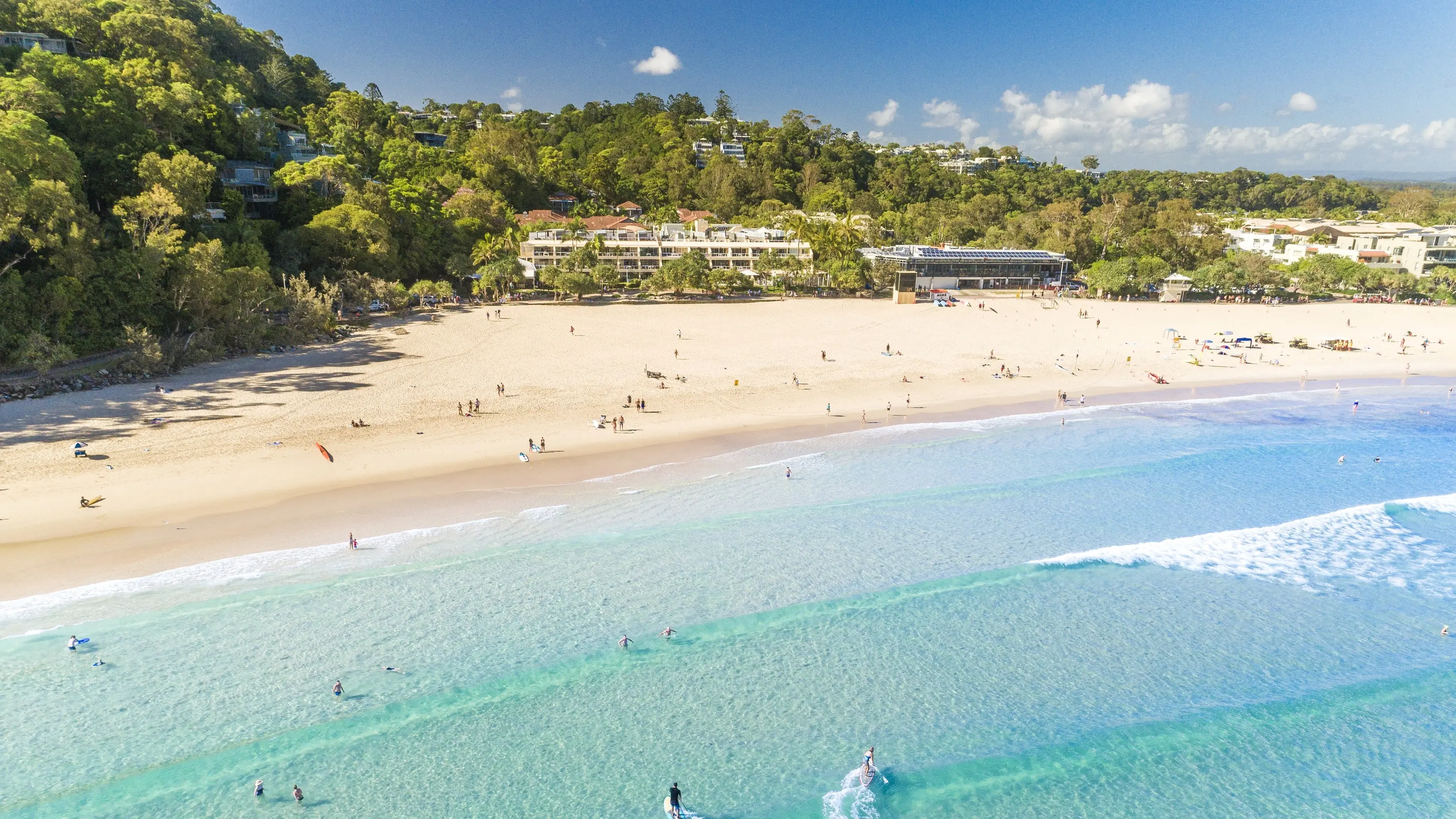 Aerial shot of wide beach with bright blue water, peach sand and fringed with trees and a beach resort building. Image credit: Tourism and Events Queensland