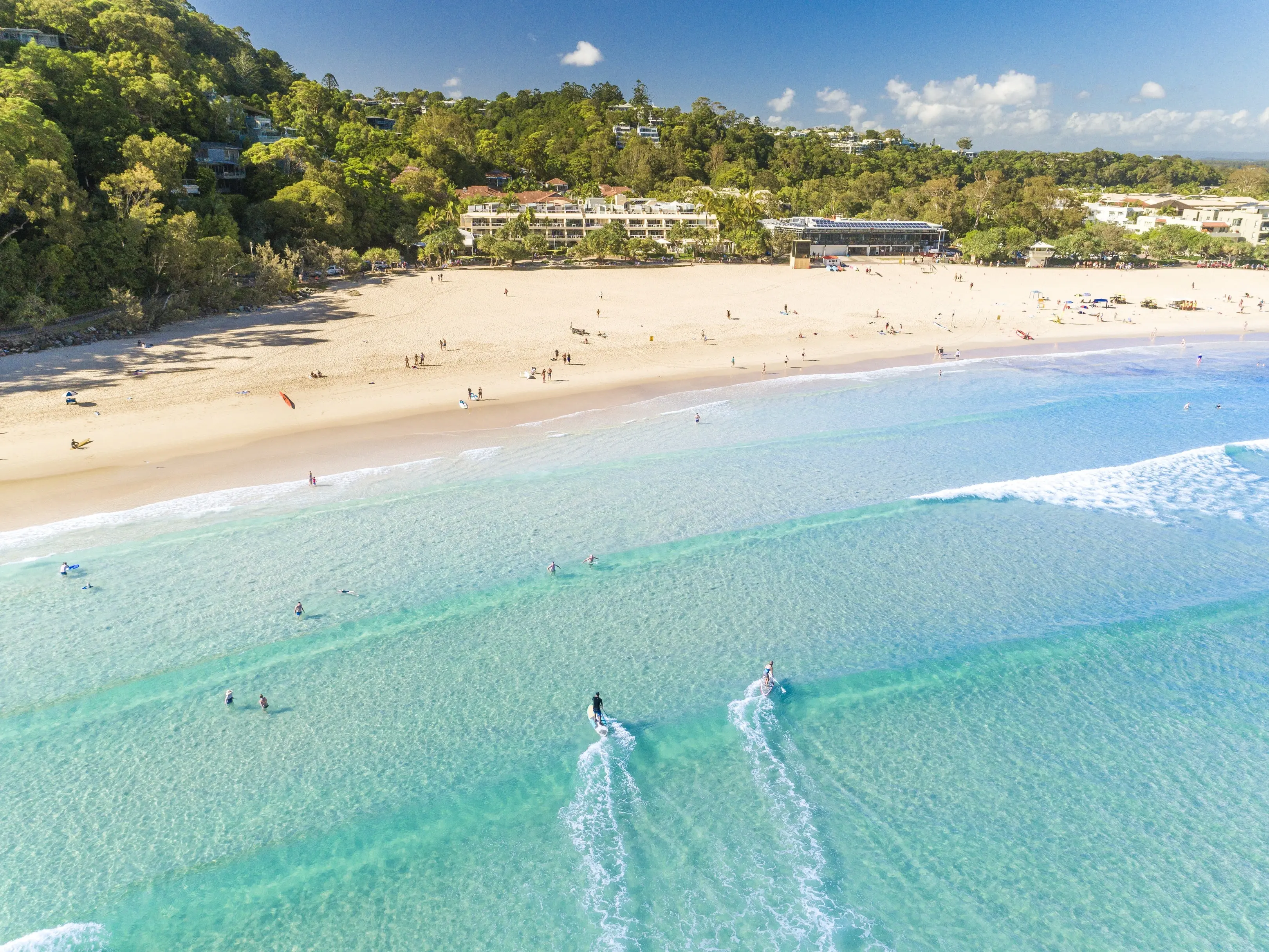 Aerial shot of wide beach with bright blue water, peach sand and fringed with trees and a beach resort building. Image credit: Tourism and Events Queensland