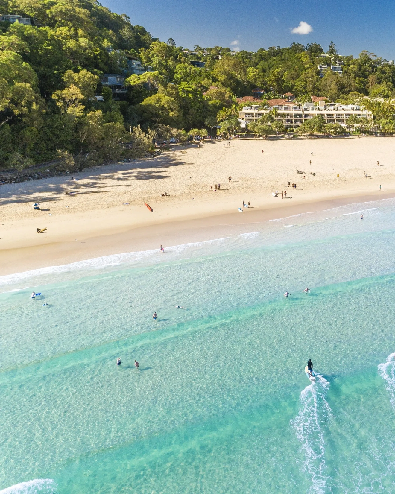 Aerial shot of wide beach with bright blue water, peach sand and fringed with trees and a beach resort building. Image credit: Tourism and Events Queensland
