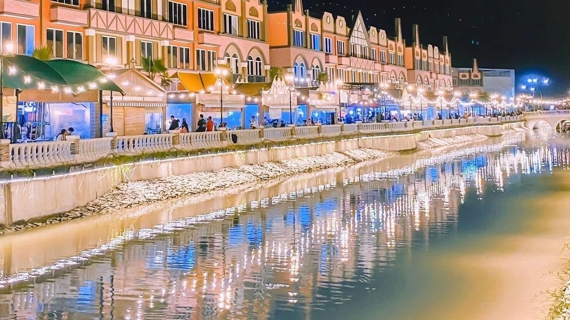 Brown buildings and outdoor dining areas along the river’s edge decorated with festive lights at night, La Riviera PIK 2, Jakarta. Image credit: @monawiskal