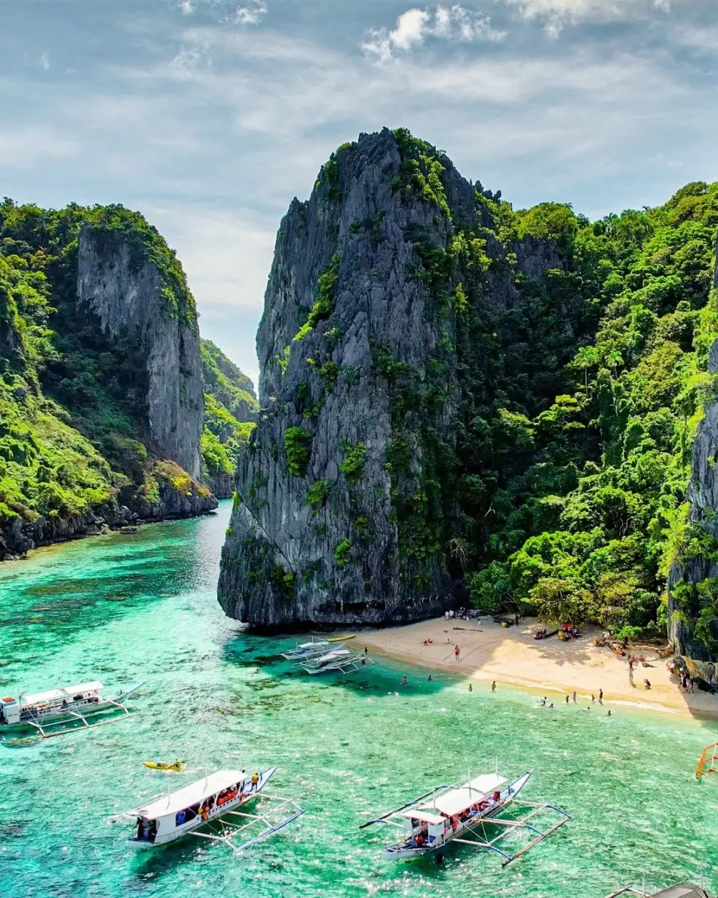 Aerial view of jagged limestone cliffs partially covered with greenery flanking a white-sand beach, with people and boats in clear, aquamarine water at El Nido, Palawa, Philippines. Image credit: stock.adobe.com