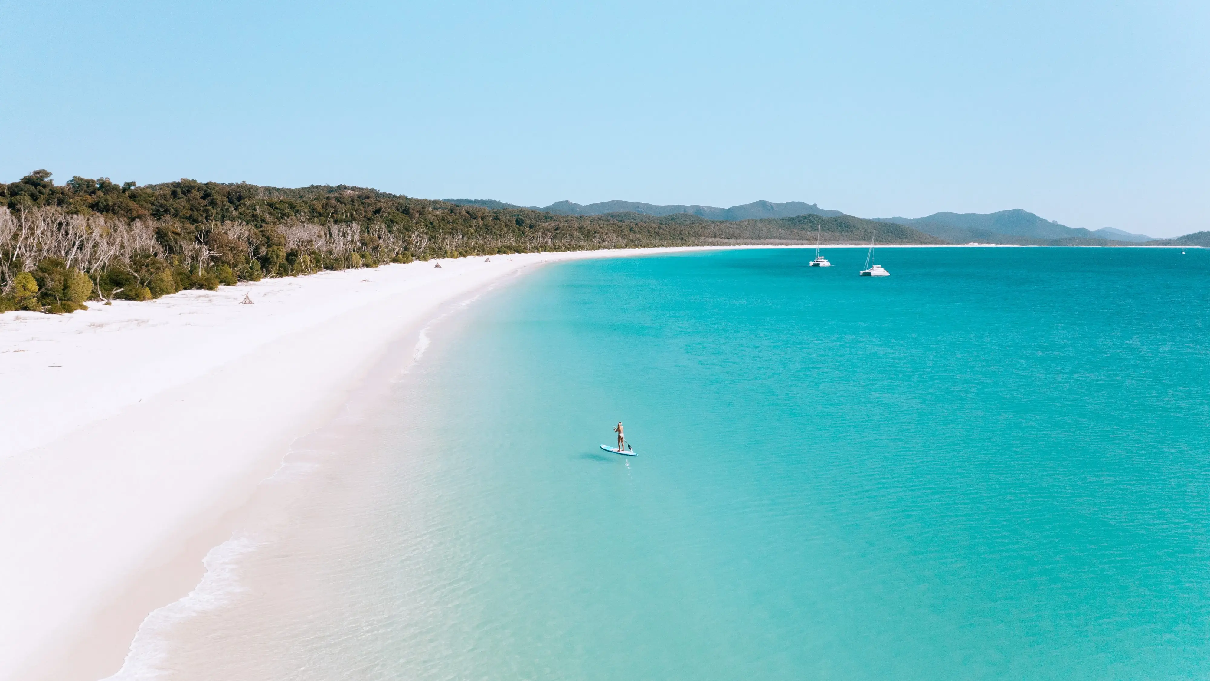 Aerial shot of white sand and bright turquoise water with paddleboarder and boats at Whitehaven Beach. Image credit: Tourism and Events Queensland