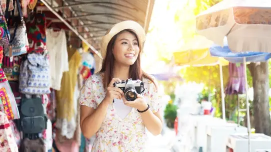 Smiling young Asian woman walks through market with camera.