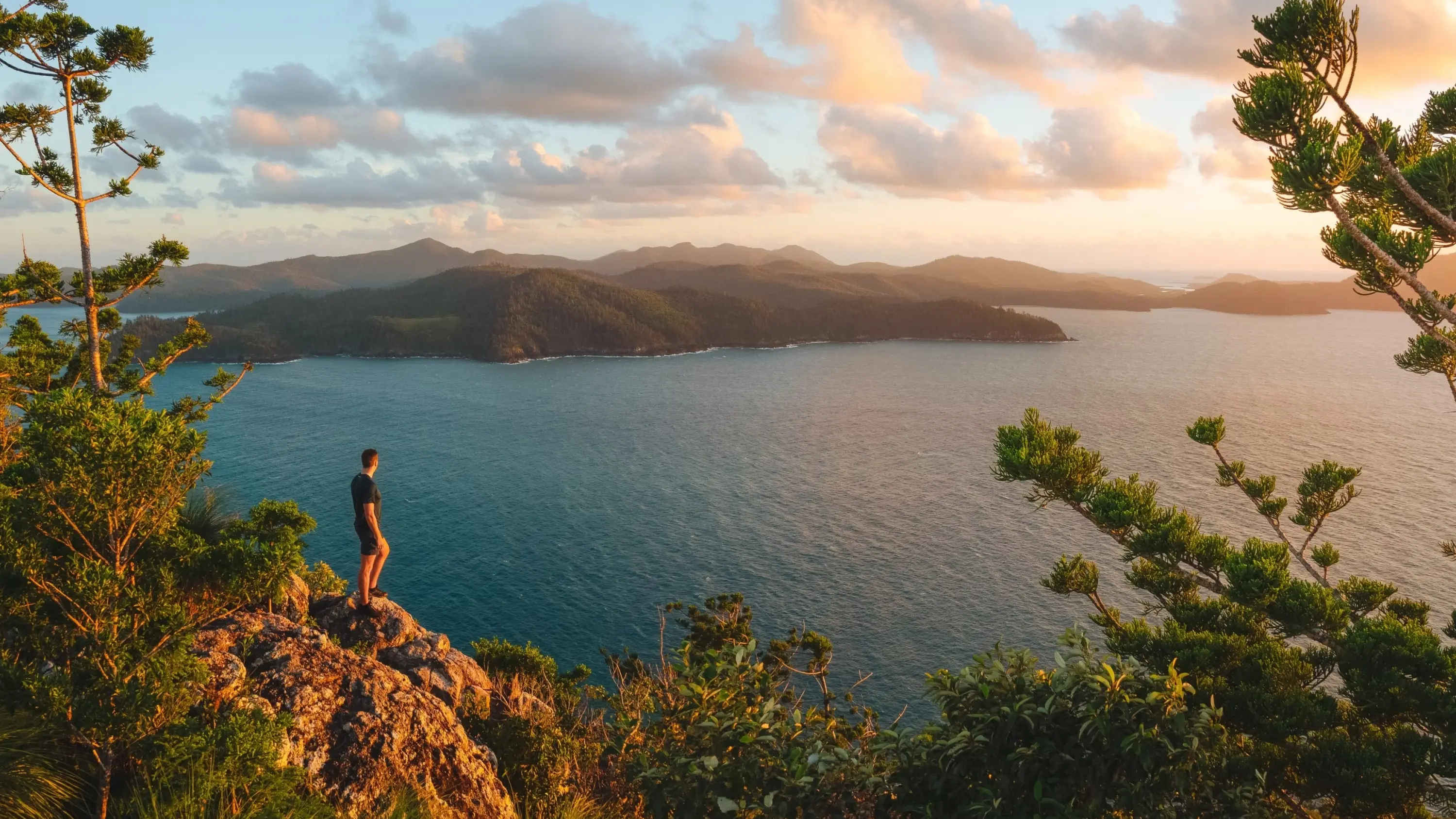 Person stands on a rocky peak at sunset, looking out over the ocean and islands. Image credit: Tourism and Events Queensland