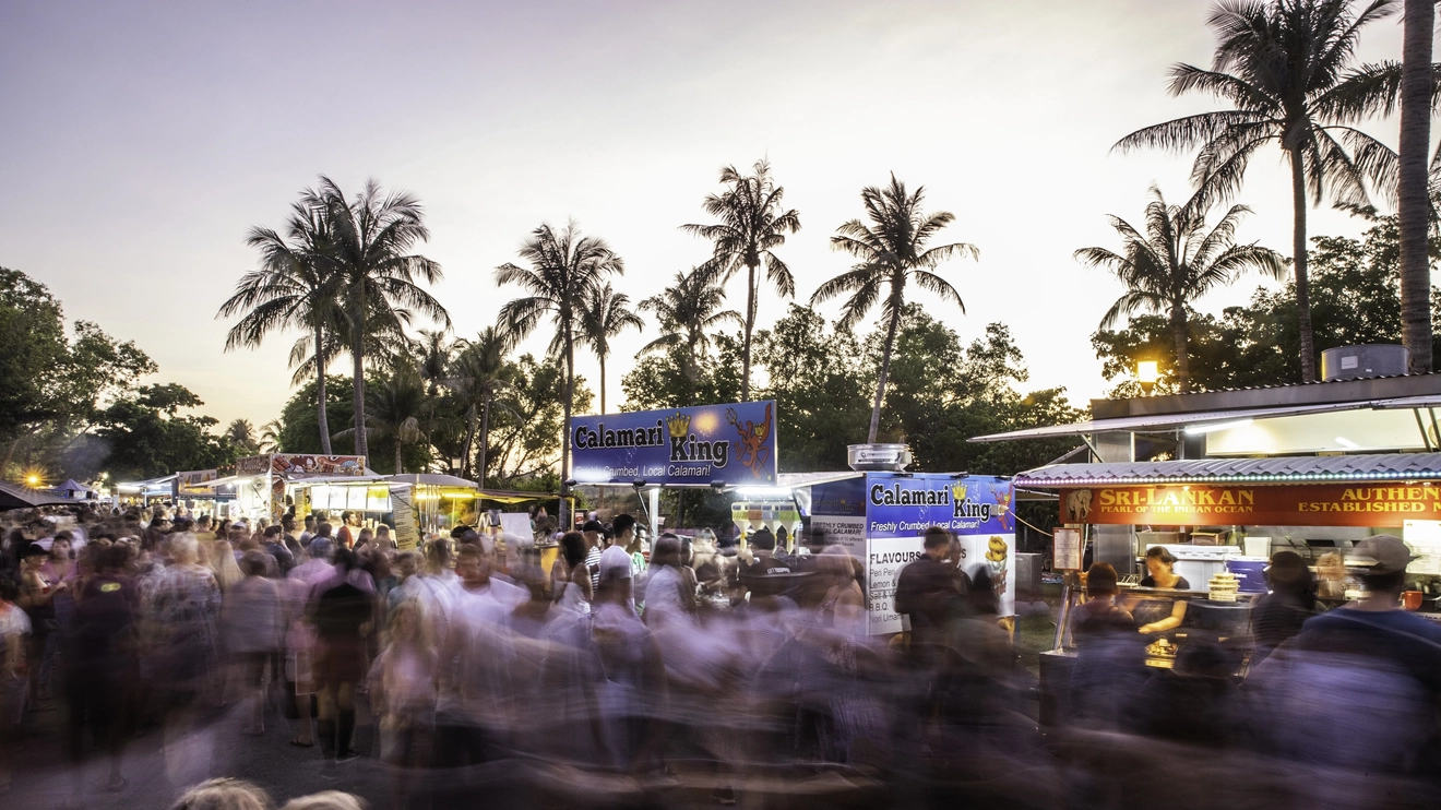 A blur of people at food stalls at Mindil Beach Sunset Market with palm trees in the background, Darwin, Northern Territory. Image credit: Tourism NT/Helen Orr