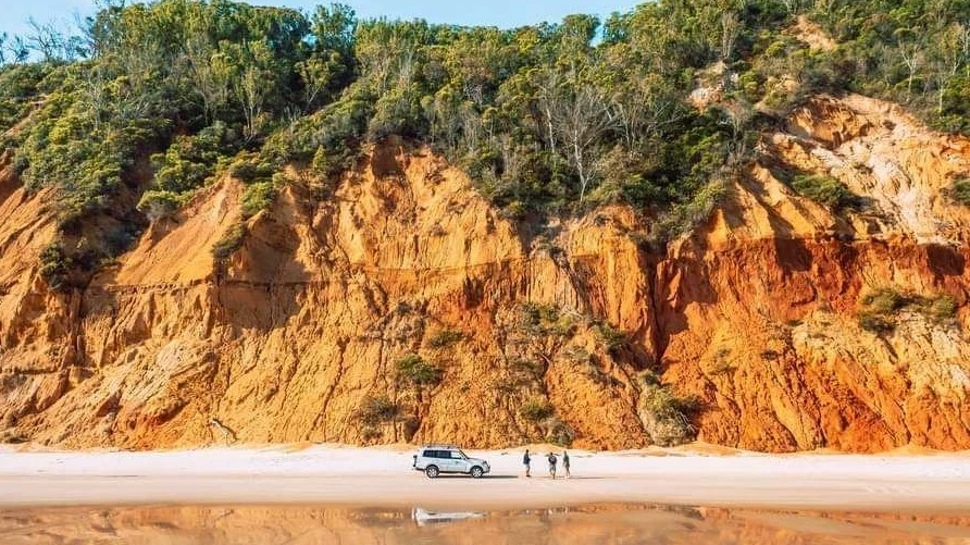 Three people and a 4WD on the sand at Rainbow Beach with deep-ochre-coloured cliffs behind them. Image credit: Tourism and Events Queensland