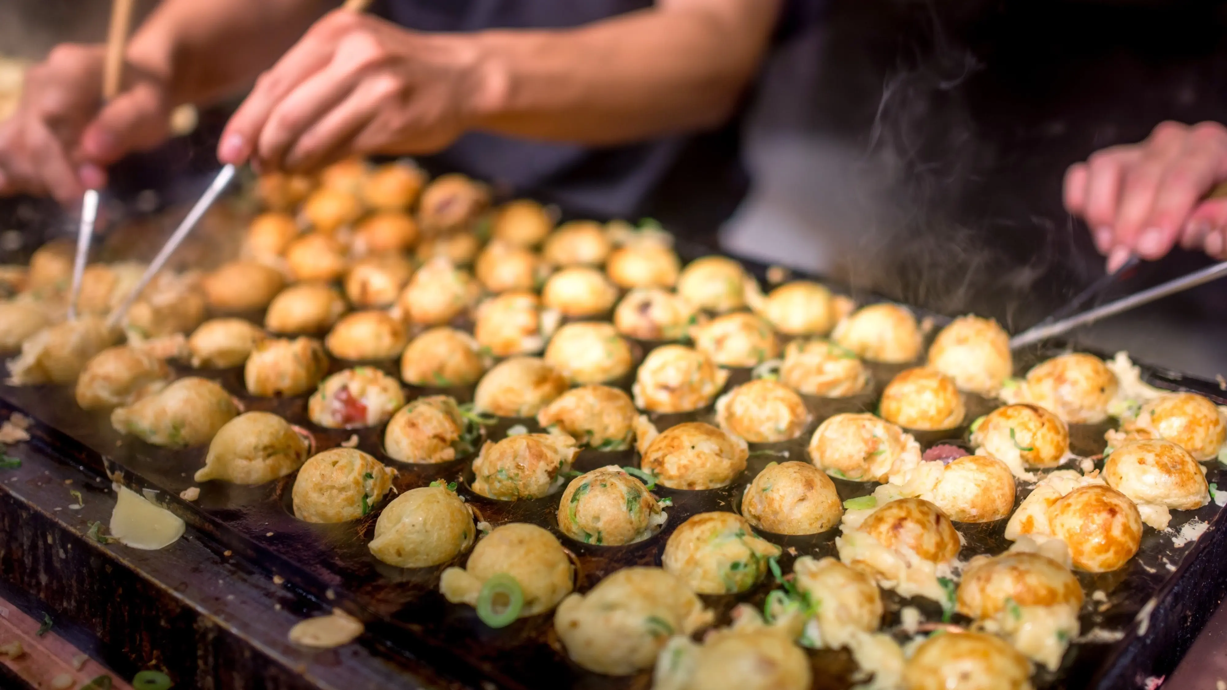 A hotplate full of cooking takoyaki octopus dumplings or balls, with a cook's hands turning them with a utensil. Image credit: stock.adobe.com