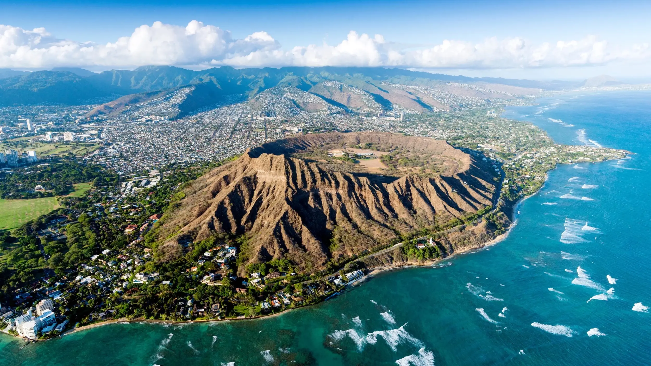 Aerial view of the Diamond Head volcanic crater, bordered by the blue water and white waves of the Pacific Ocean, with Honolulu city in the background. Image credit: stock.adobe.com