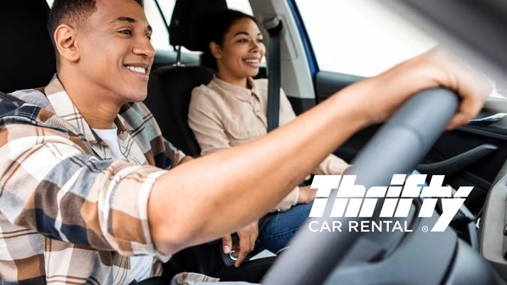 A smiling man driving a rental car with a smiling woman in the passenger seat. Thifty car rental logo is superimposed on the image.&nbsp;