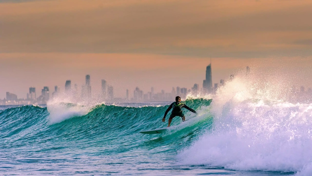A surfer rides a wave with a skyline of Surfers Paradise hotels behind it. Image credit: Shutterstock