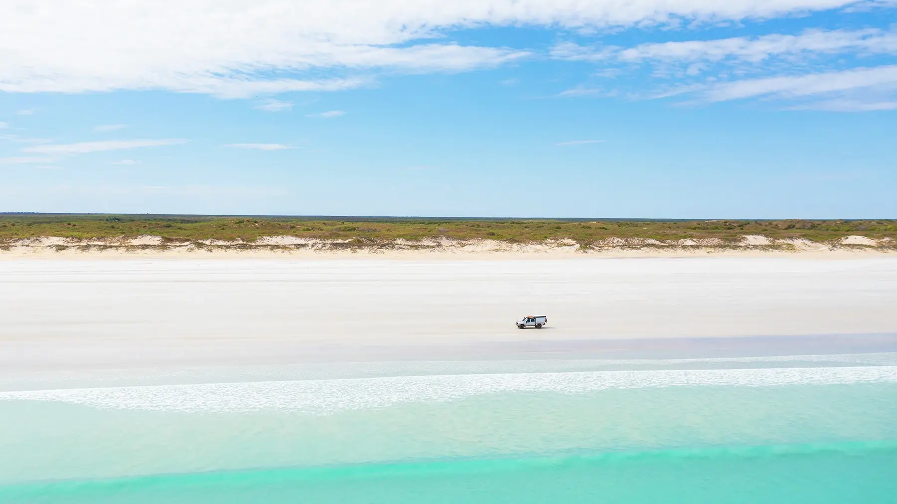 An SUV drives along the white sand of Cable Beach, Broome, with aqua-blue ocean in the foreground. Image credit: Tourism Western Australia