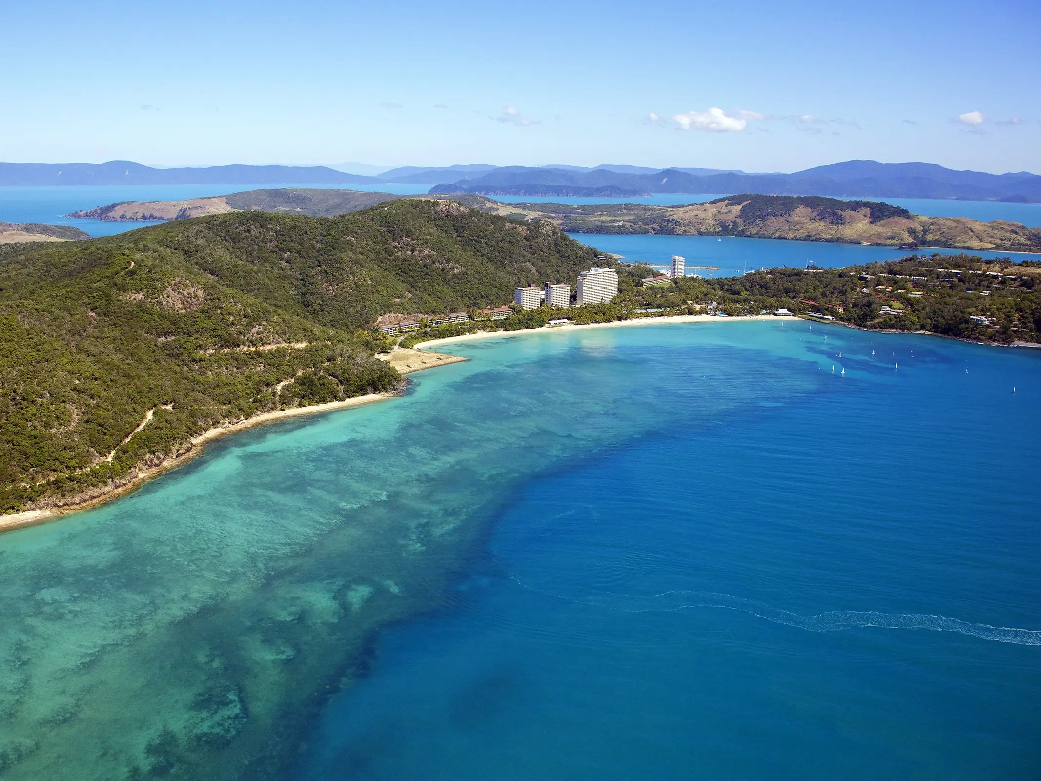 Aerial shot of foliage-covered Hamilton Island fringed with beaches and resorts, surrounded by bright blue water. Image credit: Shutterstock