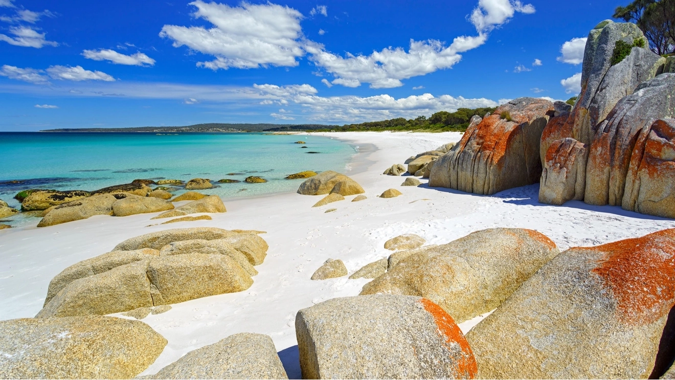Granite boulders on white sand beach with turquoise water and trees in the background, Bay of Fires.