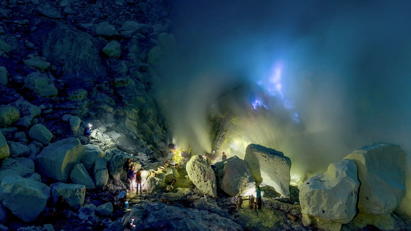 Tourists and sulfur miners standing in the Ijen Crater looking at the blue flames of burning sulfur emerging from below. Image credit: stock.adobe.com