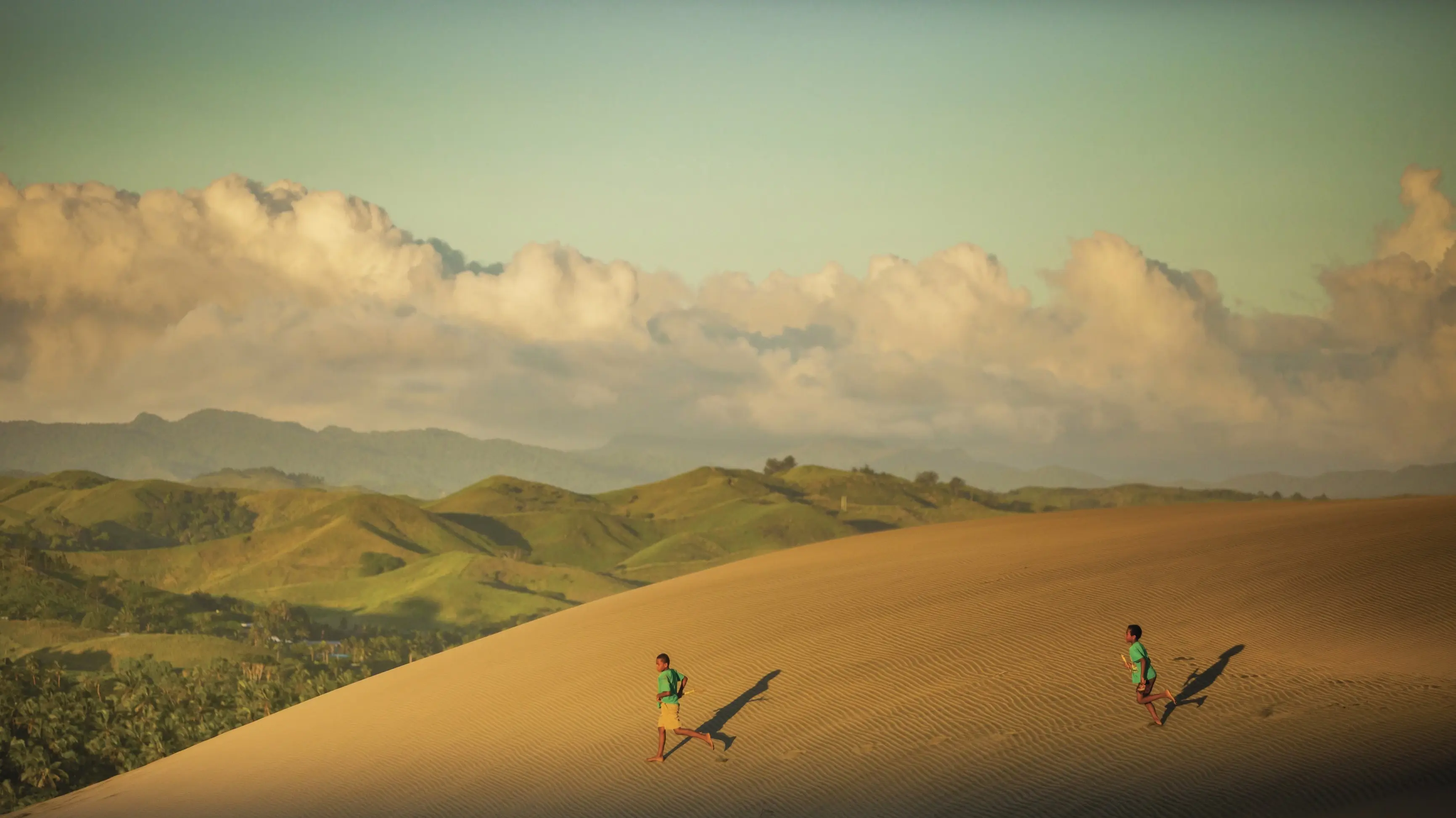 Two children running across a sand dune in Sigatoka Sand Dunes National Park, with green hills in the background. Image credit: Tourism Fiji