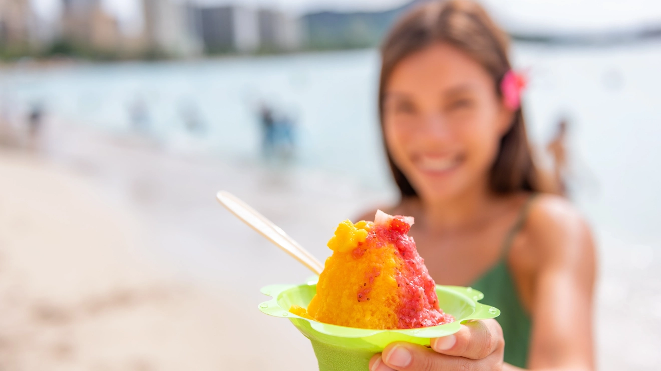 A person holds a green plastic bowl of Shave Ice, drenched in strawberry and mango puree, at Waikiki Beach, Honolulu, Hawaii. Image credit: stock.adobe.com