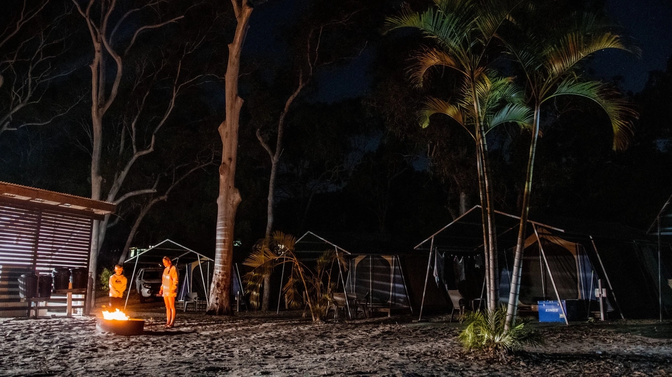 Family standing around a campfire near their tent at night, at Cathedrals on Fraser campground, K'gari. Image credit: Tourism and Events Queensland