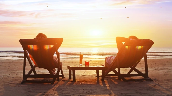 Two people relaxing on beach chairs at sunset, with a table in front holding two drinks.