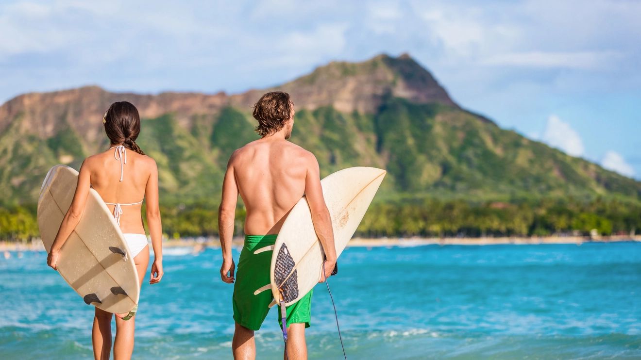 Back view of two people in bathers, carrying surfboards and looking at the water at Waikiki Beach, with Diamond Head in the background, Honolulu, Hawaii. Image credit: stock.adobe.com