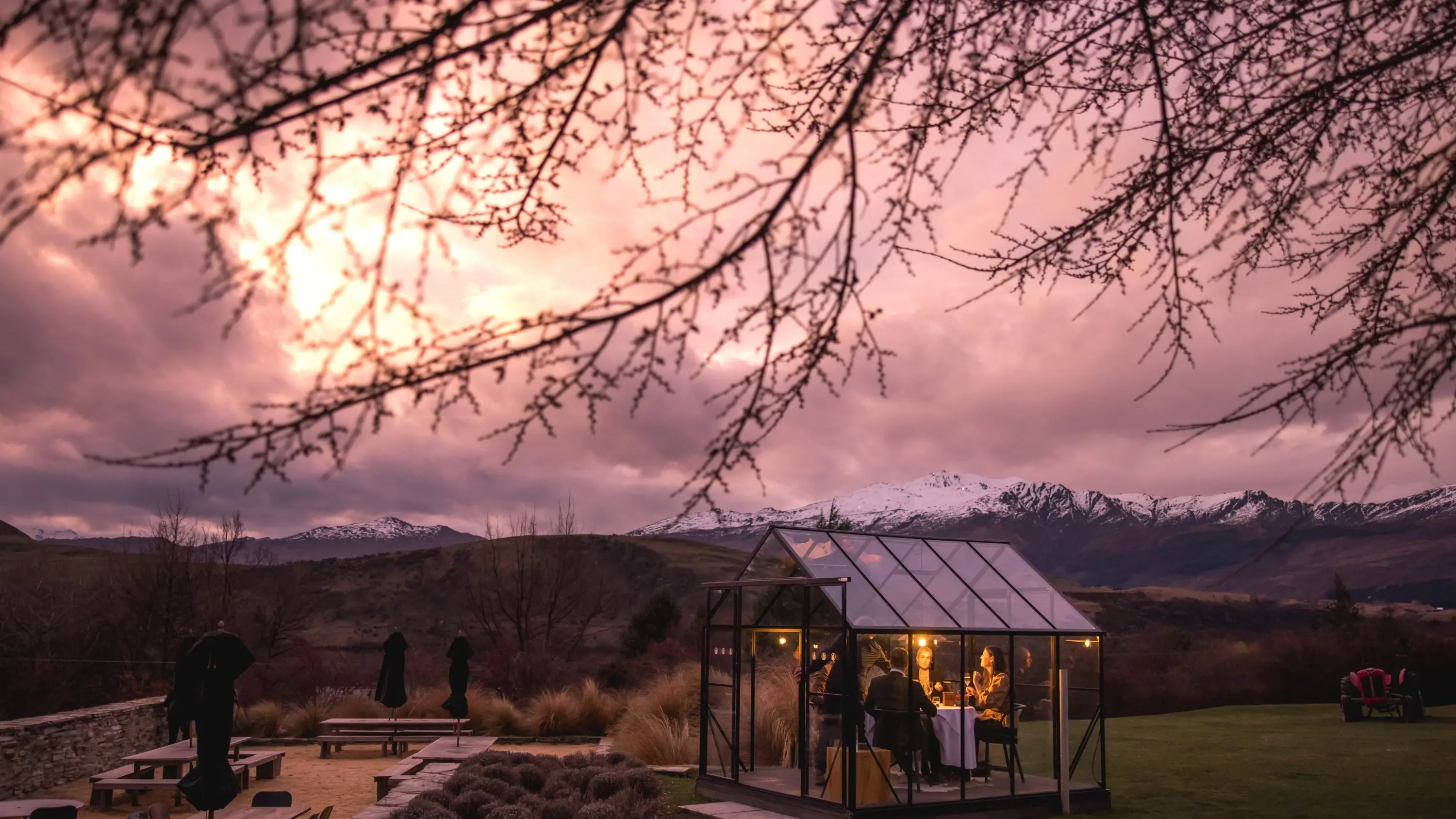 A small group of people in a glass conversatory on the lawn at dusk, with snow-capped mountains in the background, at Amisfield Restaurant and Cellar Door, Lake Hayes, Queenstown. Image credit: Tourism NZ/Miles Holden