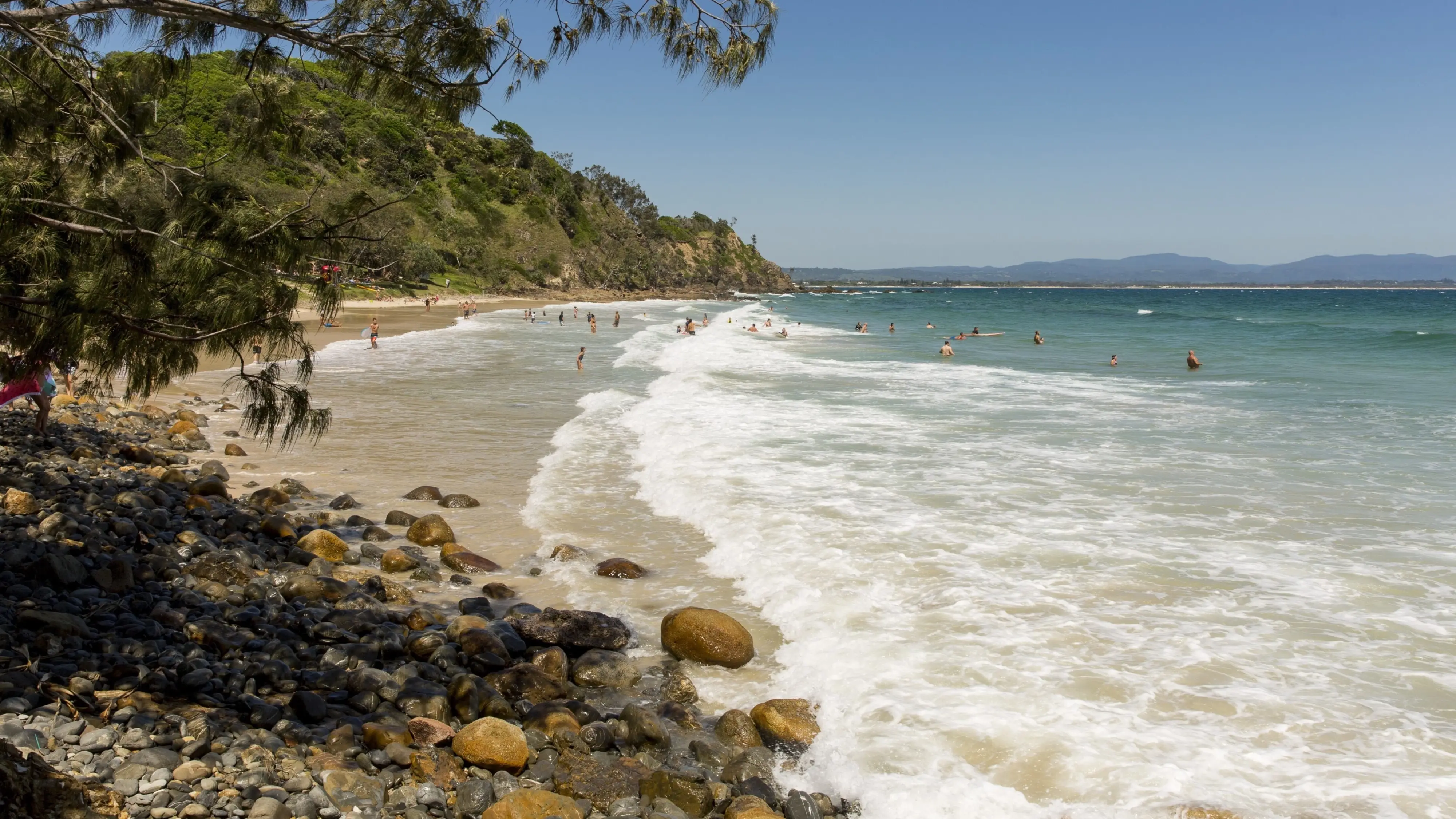 Wategos Beach, with trees and rocks in foreground, curved shore and gentle waves. Image credit: Destination NSW