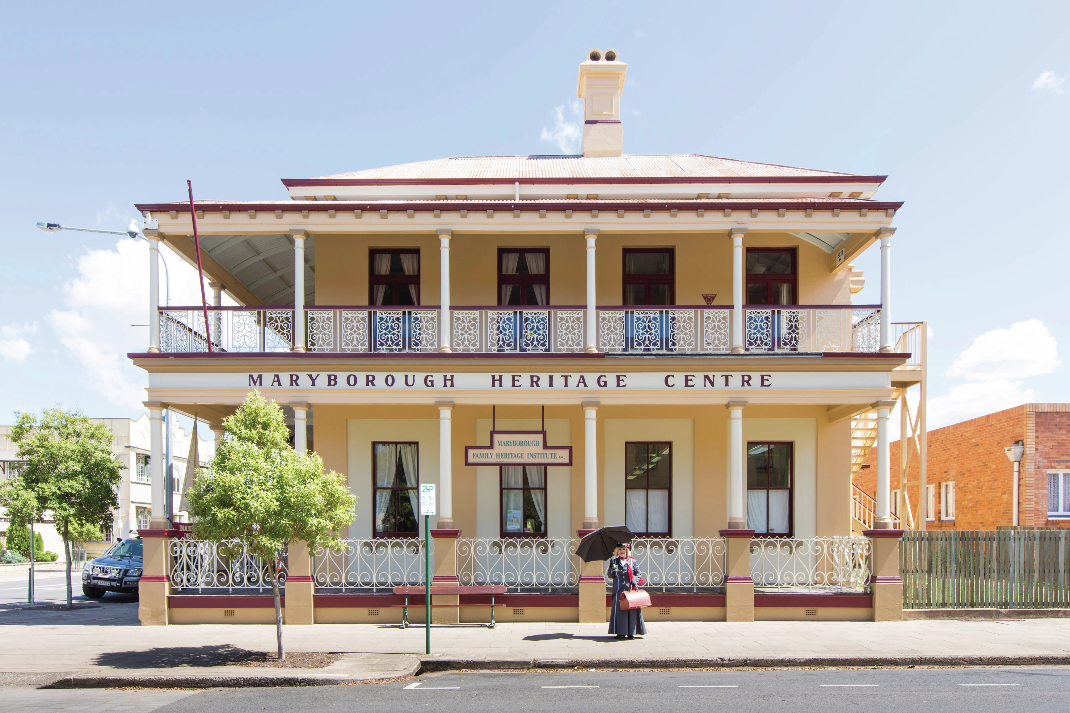A woman in historic clothing stands outside the ornate, double-story Maryborough Heritage Centre on a sunny day. Image credit: Andrew Tallon/Tourism and Events Queensland