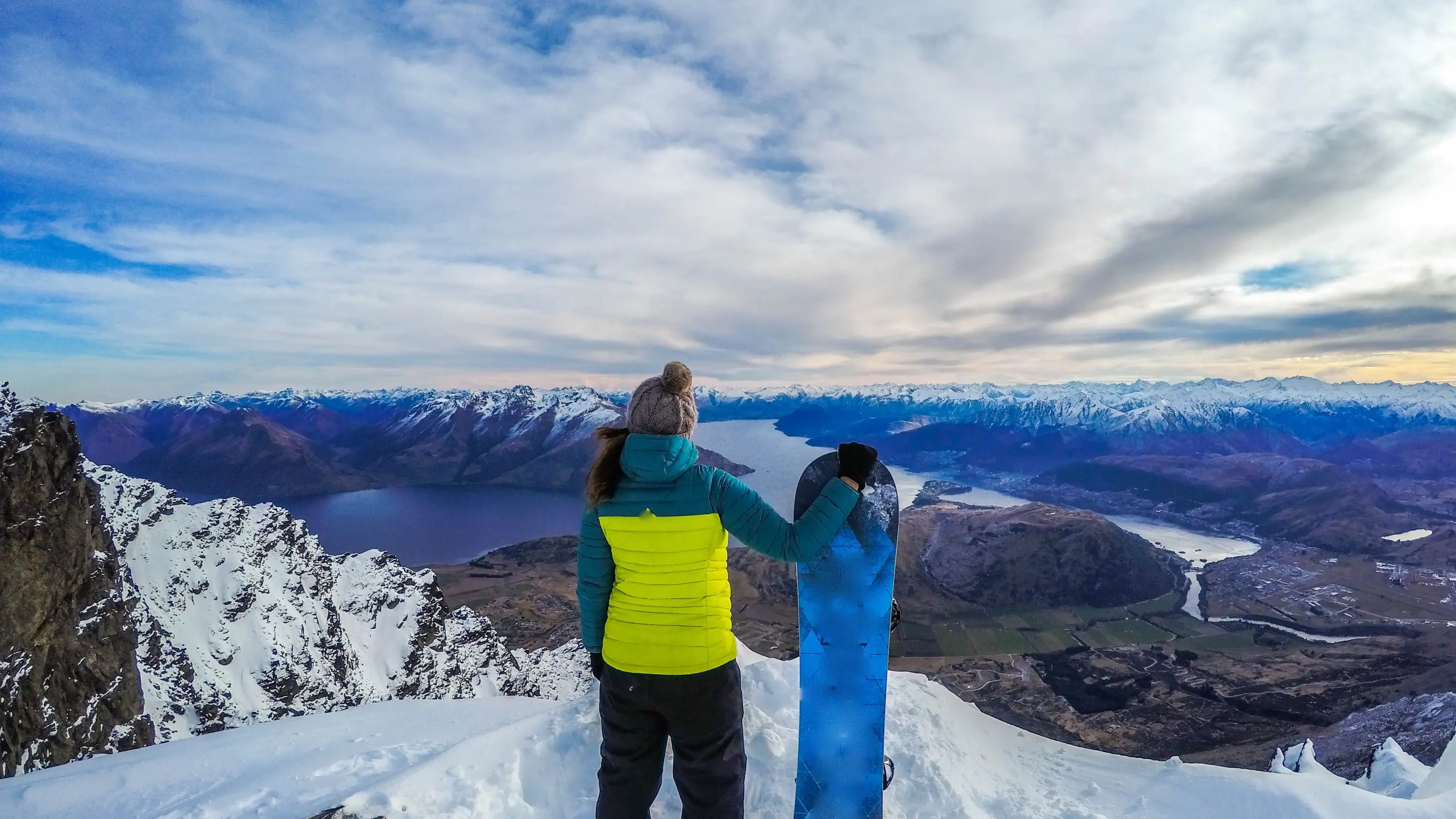 Woman with snowboard stands on a ski slope looking at Lake Wakatipu and snow-covered mountains in the distance, Queenstown, New Zealand. Image credit: stock.adobe.com