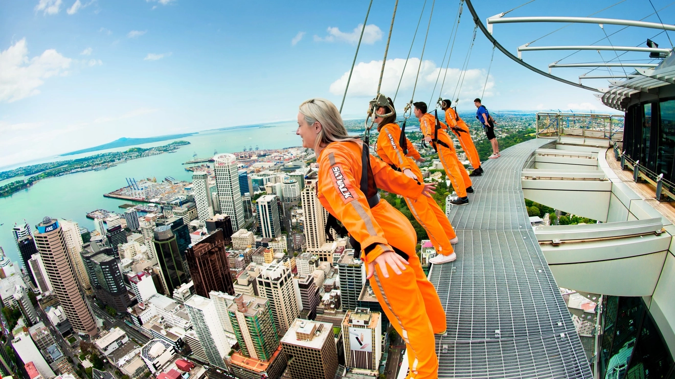 People in orange safety suits and rigging lean over the edge of the SKYWALK platform at SkyTower, Auckland, New Zealand. Image credit: Auckland Brand Library