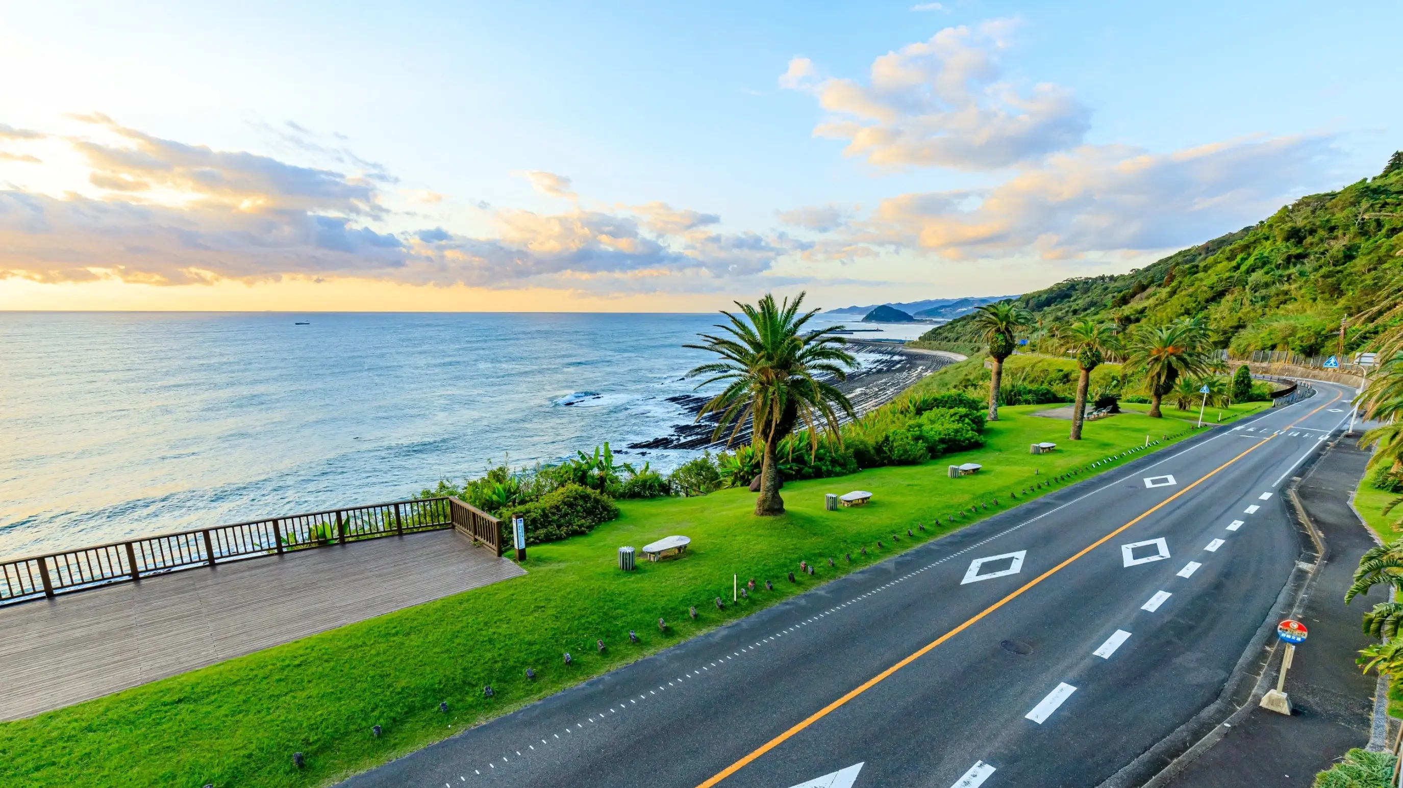 Nichinan coastline at sunrise, with road, palm trees and grass in the foreground and ocean, coast and hills in the background, as seen from Phoenix, Miyazaki. Image credit: stock.adobe.com