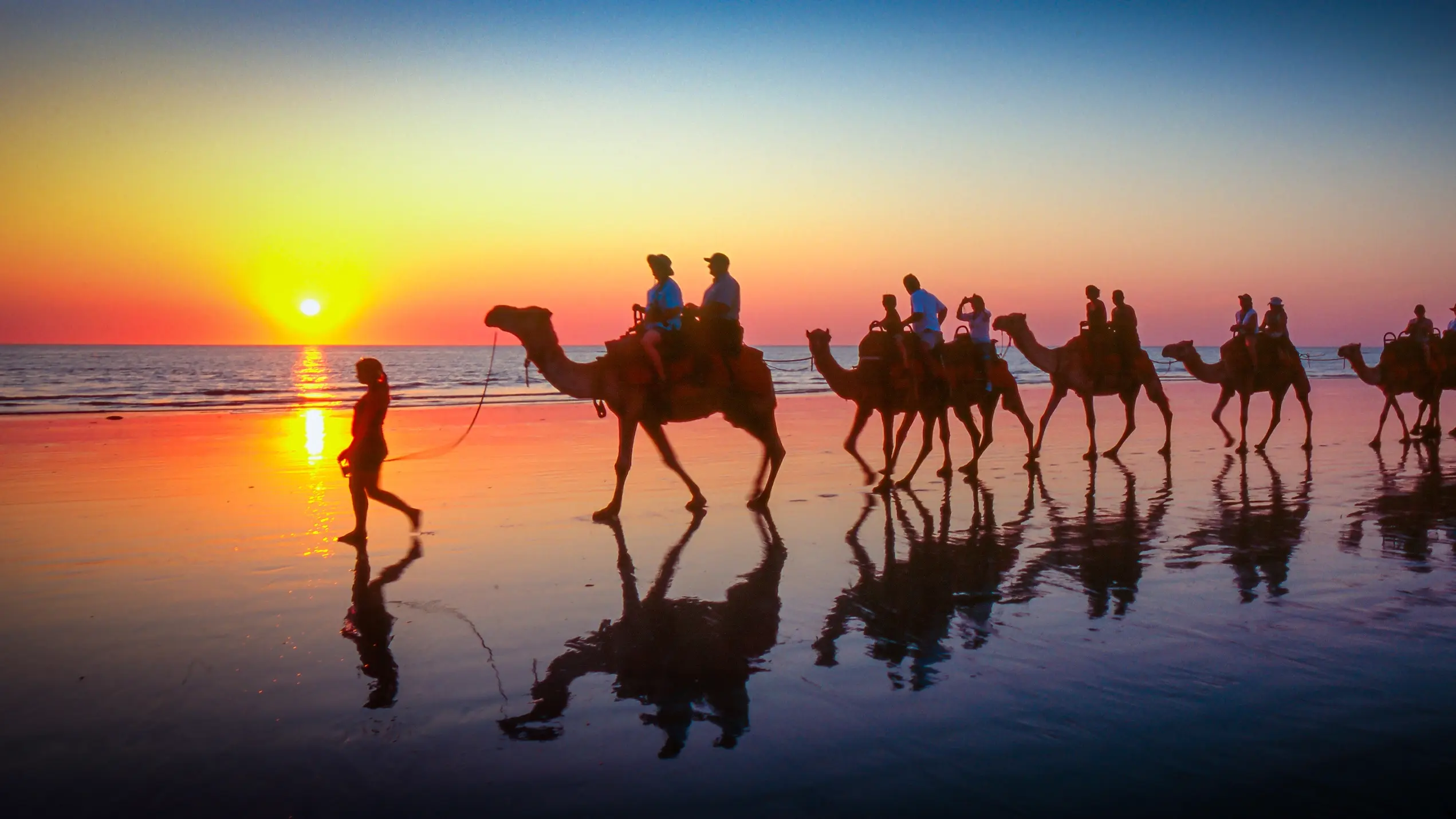 Silhouetted against a sunset sky, a woman leads a group of tourists riding camels along Cable Beach. Image credit: stock.adobe.com