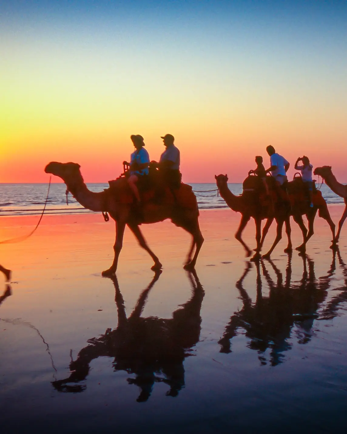 Silhouetted against a sunset sky, a woman leads a group of tourists riding camels along Cable Beach. Image credit: stock.adobe.com