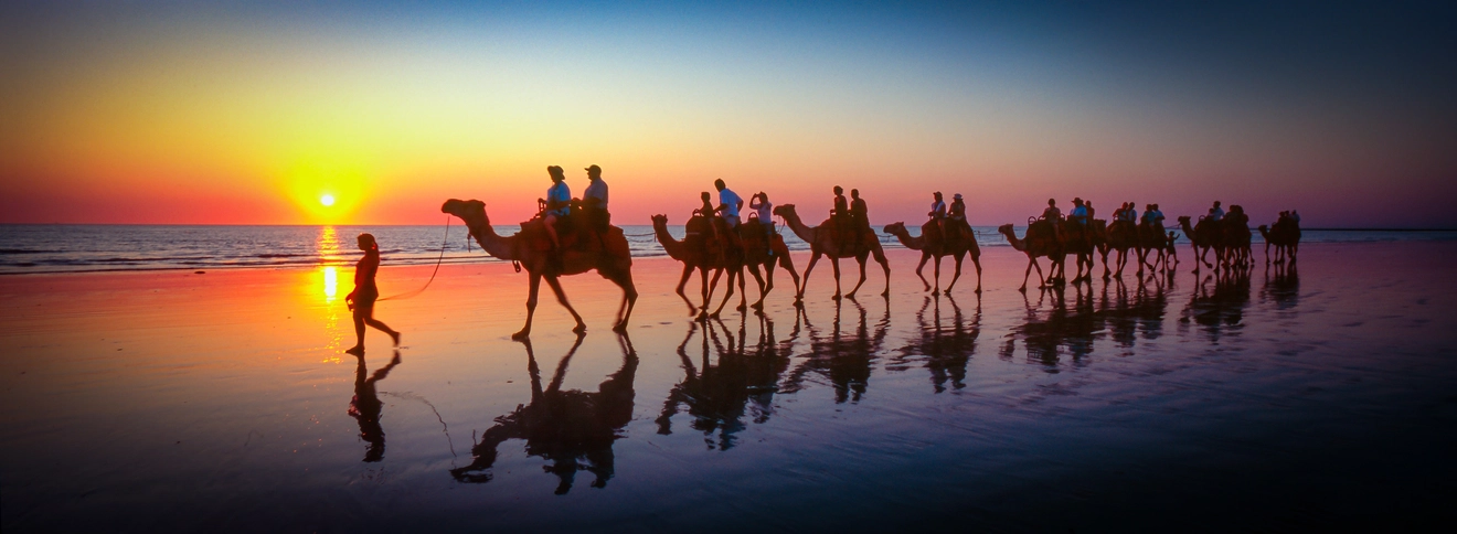 Silhouetted against a sunset sky, a woman leads a group of tourists riding camels along Cable Beach. Image credit: stock.adobe.com