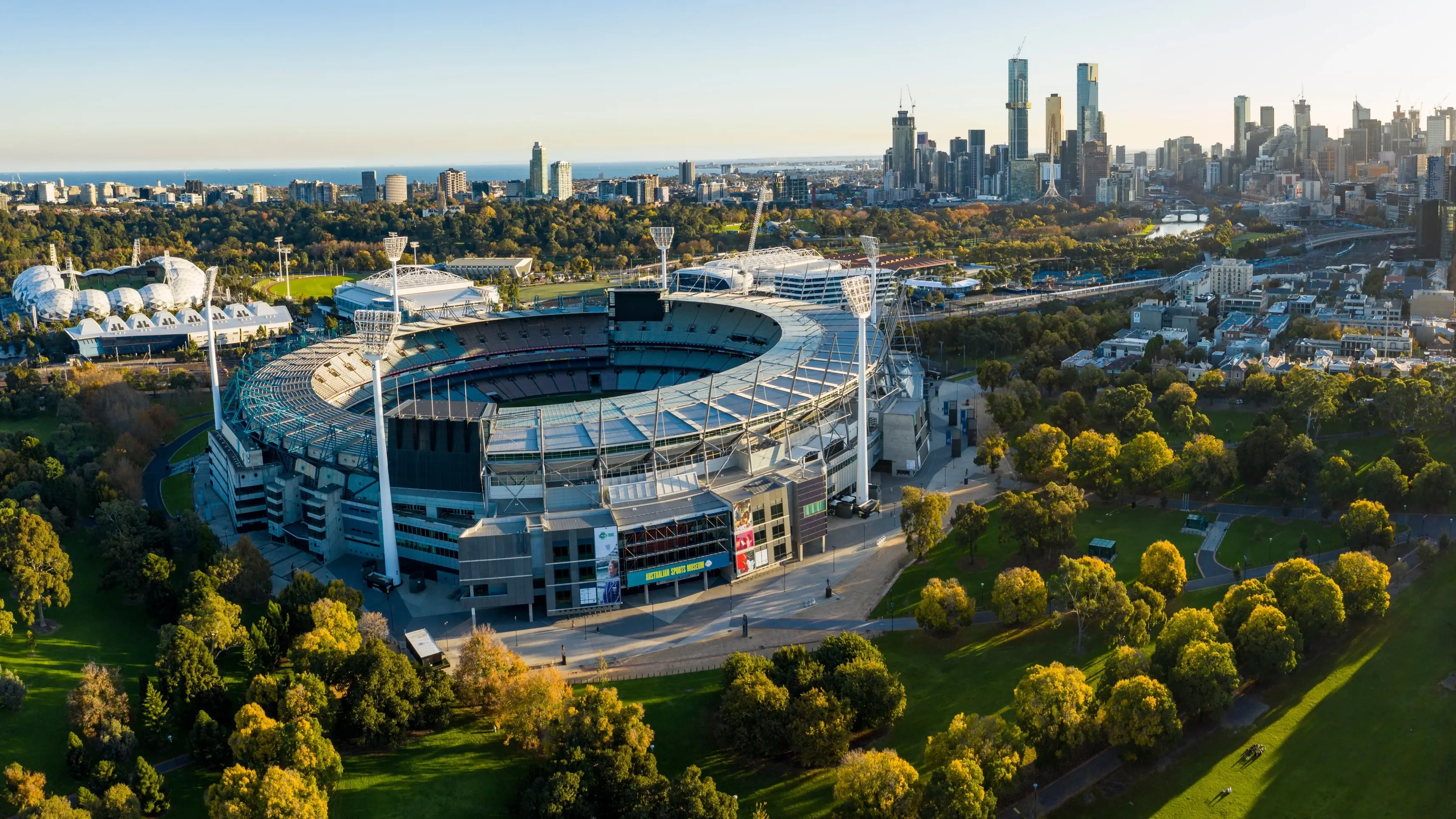 Aerial view of Melbourne Cricket Ground on a sunny day, surrounded by trees with the city skyline in the background. Image credit: Michael Evans/stock.adobe.com