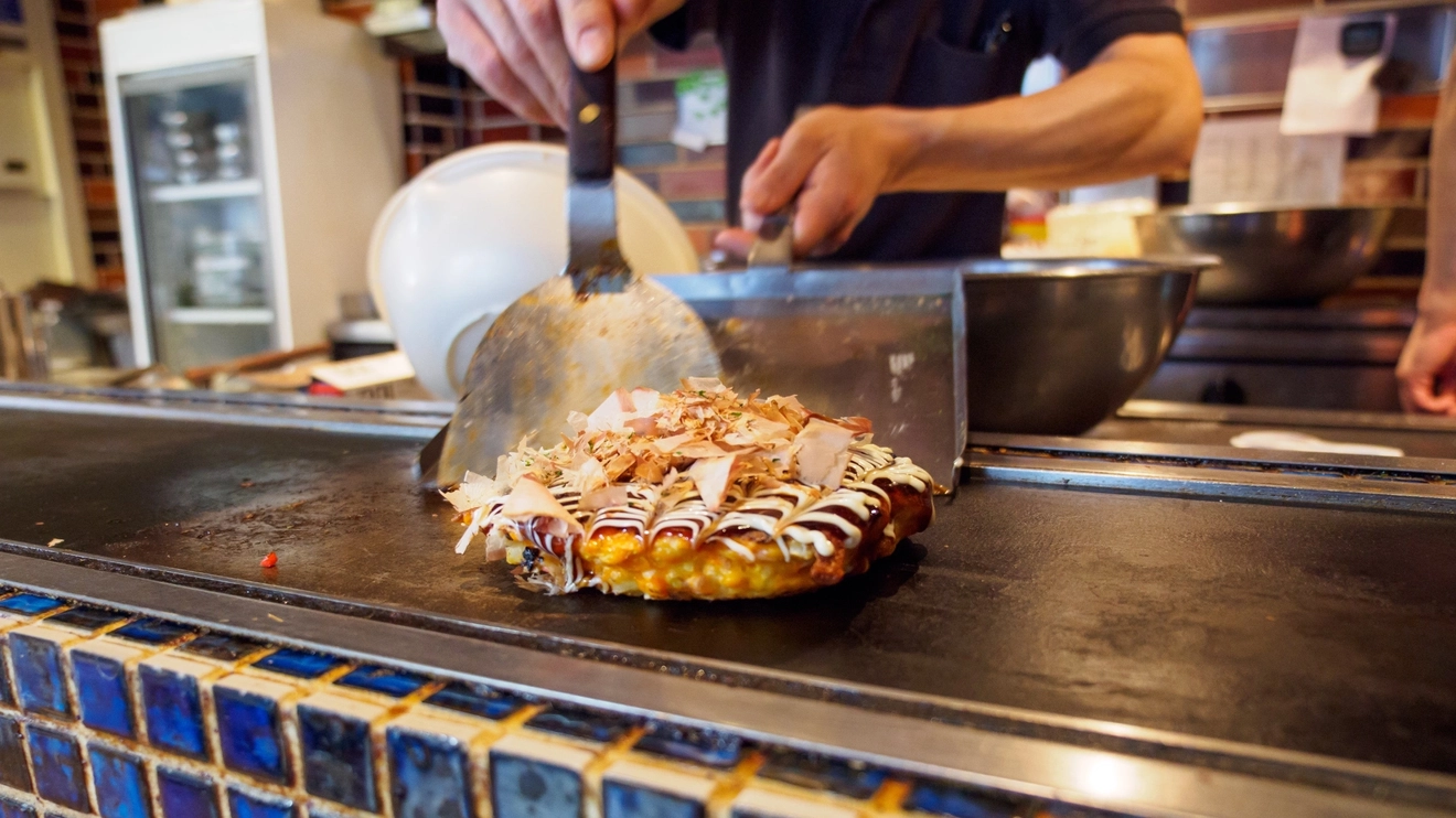 Close-up of an okonomiyaki cooked on a teppan grill, topped with Katsuobushi fish flakes, sauce, and mayonnaise. Dotonbori, Osaka, Japan. Image credit: stock.adobe.com