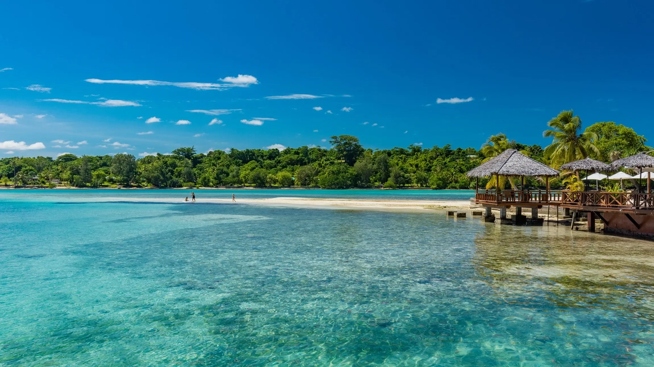 Thatched shacks over calm turquoise water, with palm trees and a sandbar in the background, Erakor Island, Efate, Vanuatu. Image credit: stock.adobe.com