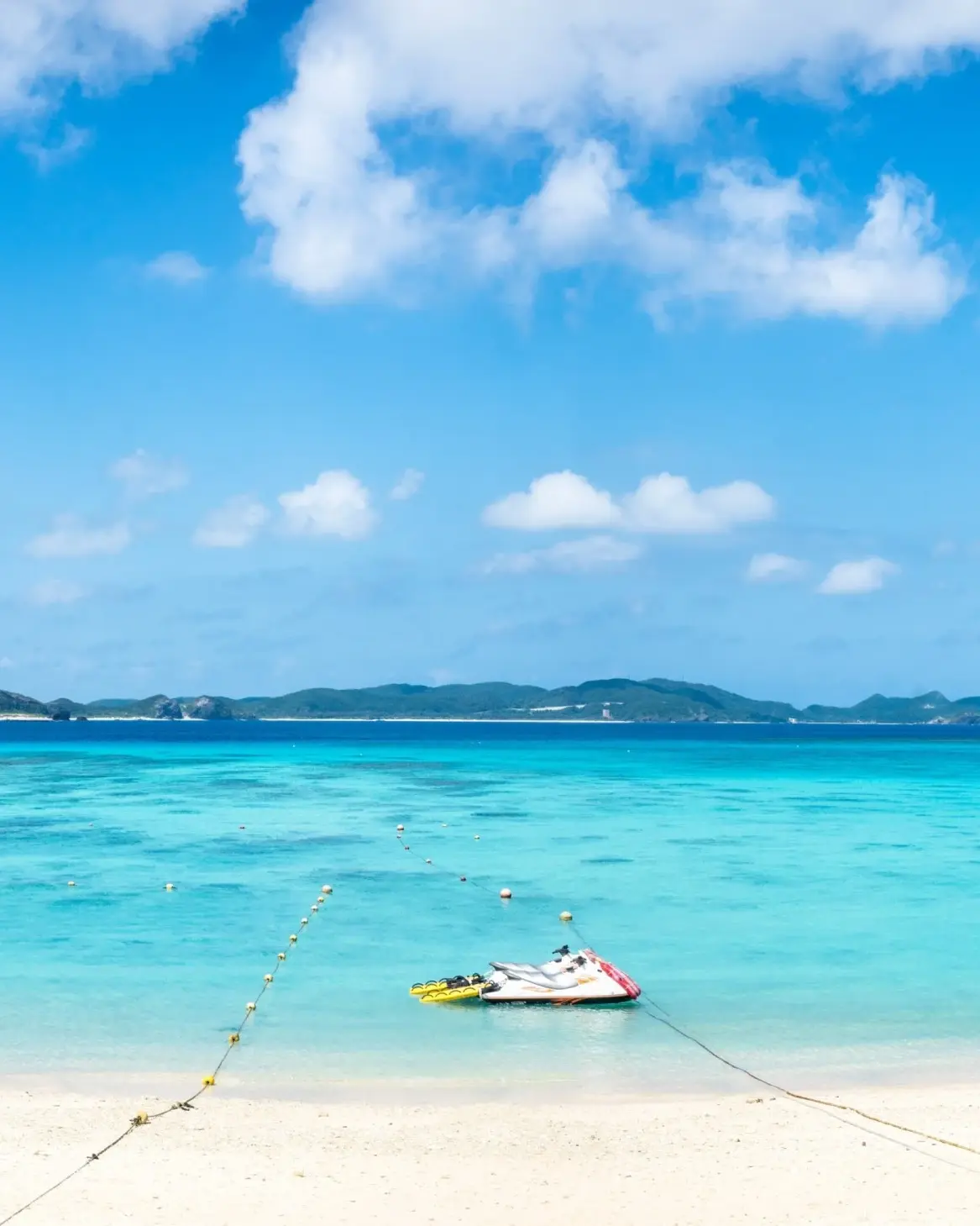 White-sand beach with jetskis and canoe moored in turquoise water, green hill on shore and islands on the horizon, at Tokashiku Beach, Tokashiku Island, Okinawa, Japan. Image credit: stock.adobe.com