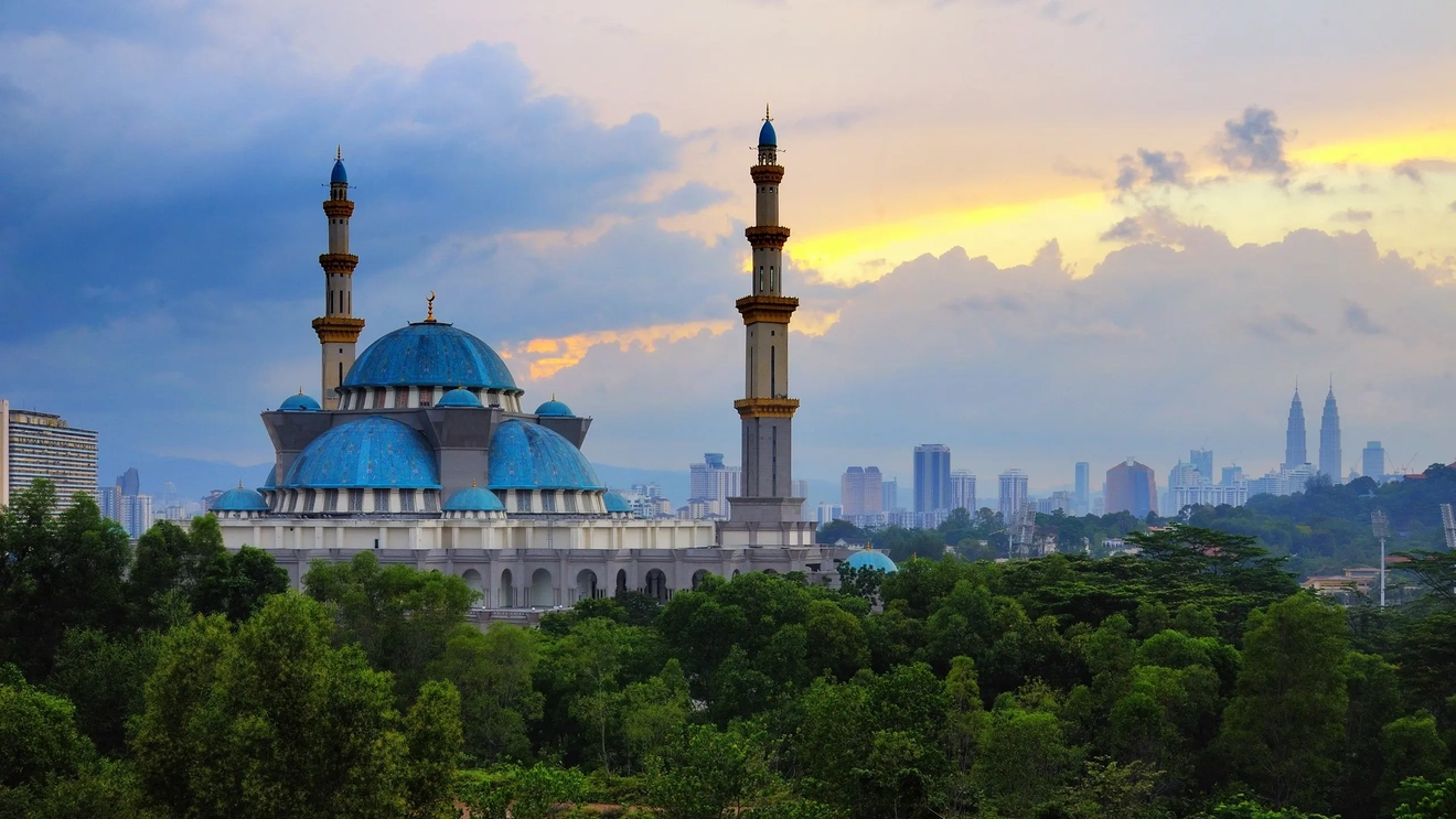 The blue domes and white pillars of the Federal Territory mosque in Kuala Lumpur, Malaysia, at sunrise, with trees in the foreground. Image credit: Shutterstock