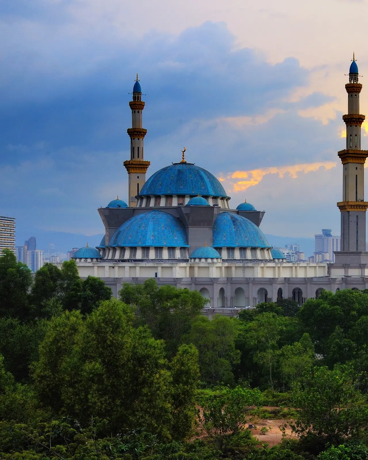 The blue domes and white pillars of the Federal Territory mosque in Kuala Lumpur, Malaysia, at sunrise, with trees in the foreground. Image credit: Shutterstock