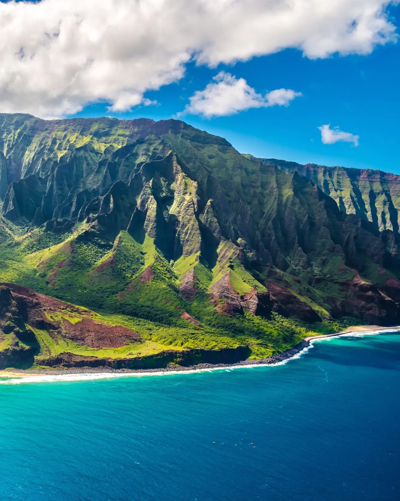 Aerial view of rocky, green volcanic cliffs bordered by white sand beach and aquamarine water on the Na Pali Coast, Kauai, Hawaii. Image credit: stock.adobe.com