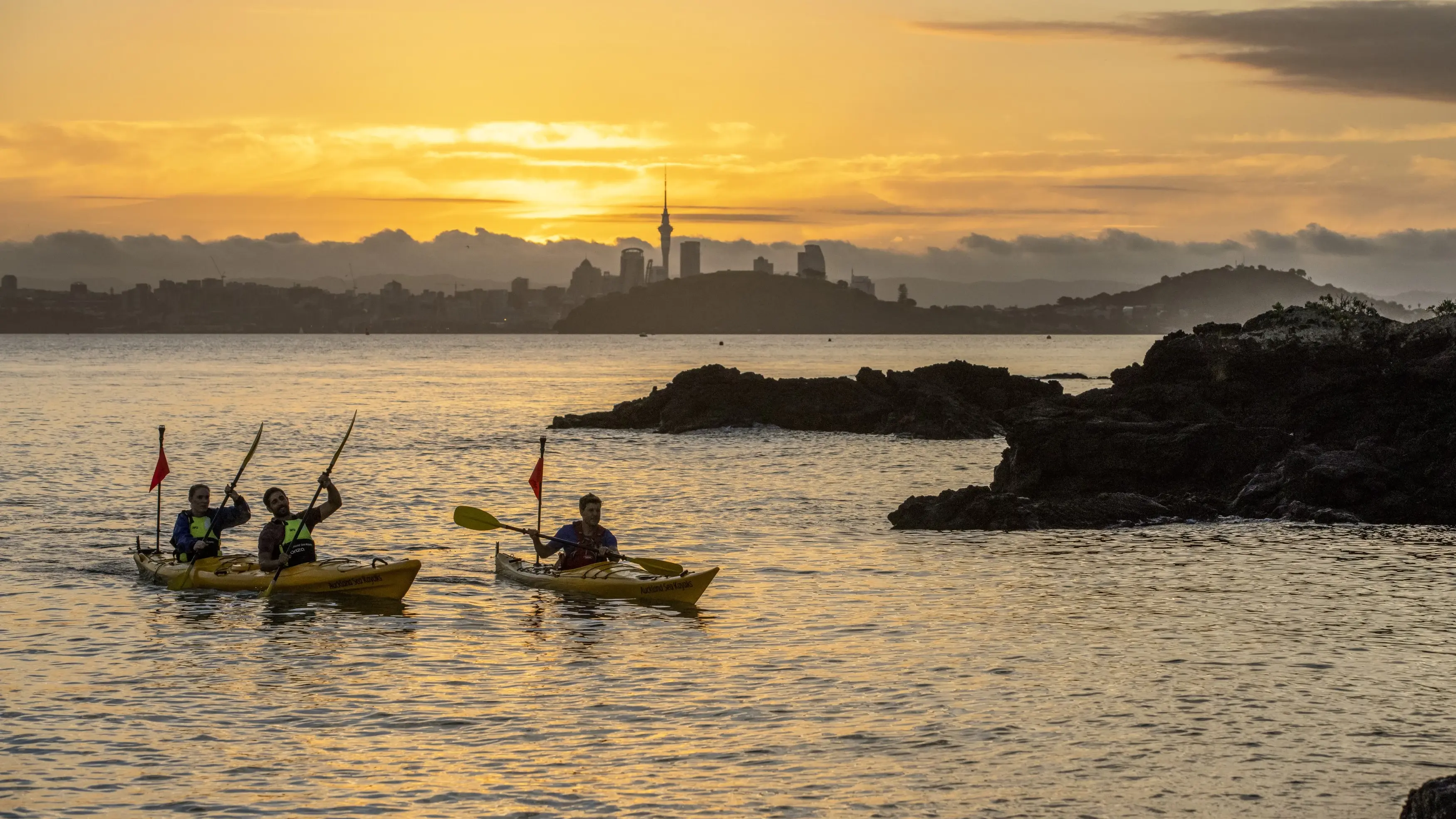 Kayakers in a rocky bay at sunset with cityscape in background, Auckland, New Zealand. Image credit: Tourism NZ/Miles Holden